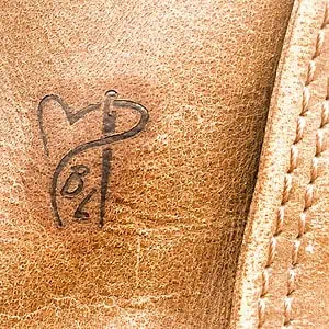 Auf diesem Foto kann man einen Brandestempelabdruck betrachten, der die Initialen B und L enthält. Die beiden Buchstaben sind von einem Garn umrahmt. Der Faden bildet ein Herz, dieses wird von einer Nadel unterbrochen. Das Motiv ist auf Leder gestempelt worden.