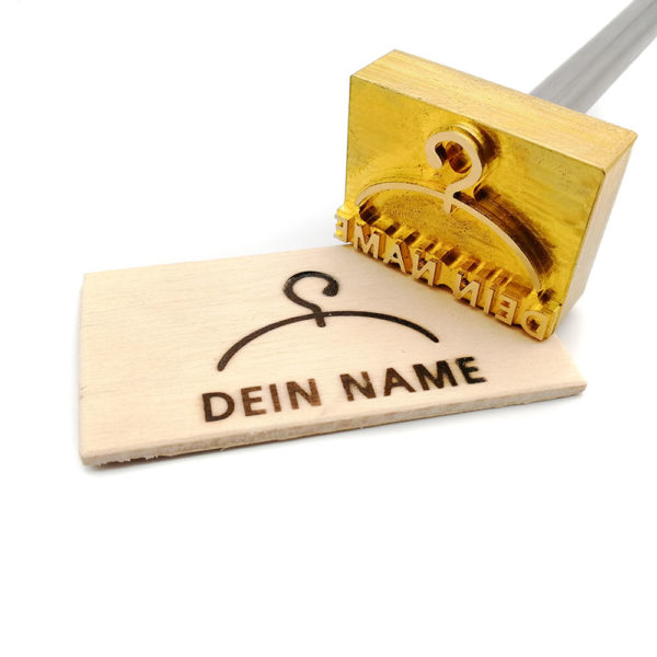 Dieser Stempel hat einen Kleiderbügel als Motiv und unterhalb davon befindet sich ein Schriftzug in großen Lettern mit „dein Name“. Der Schriftzug kann auf Kundenwünsche und in unterschiedlichen Ausführungen angepasst werden. Zu sehen ist der Stempel hierbei in der externen Ausführung als Probedruck auf einer kleinen Holzscheibe. Anbei ist das Motiv in elektrischer Ausführung fotografiert. Ein Brennkolben mit schwarzem Griff und einem Edelstahlkolben gefolgt von der Messingstück. Das Gerät liegt auf einem aufgeklappten Ständer. Ein gefräst ist das Motiv in einer Messingplatte und wurde auf ein kleines Stück Holz eingebrannt.