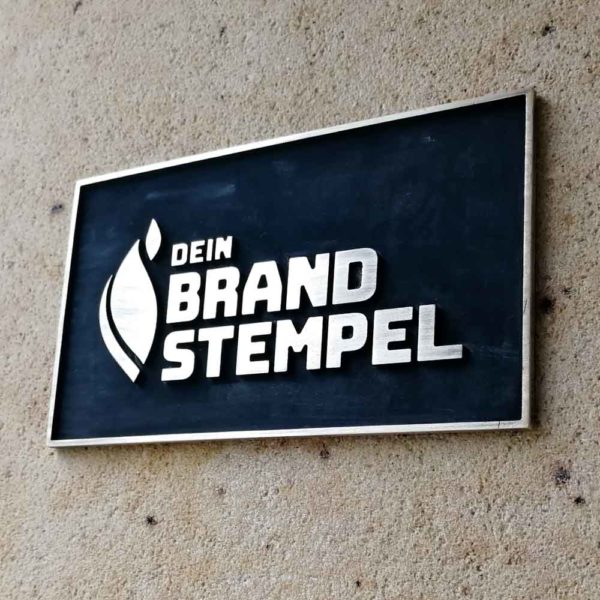 Hier ist ein Messingschild zu sehen, welches schwarz patiniert wurde. Die Schrift sticht dabei golden hervor und wird von einem Steg umrahmt. Als Motiv wurde das Logo der Firma Brandstempel verwendet.