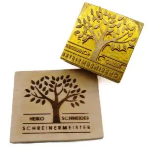 Ein stilisierter Baum mit einer Baumkrone aus der drei Äste auswachsen, welche sich dann wieder in dünnere Äste aufteilen. Der Baum steht auf einer Linie welche jeweils von einer oberen und unteren Linie unterstützt wird. In der mittleren oberen „Schiene“ findet sich der Schriftzug HEIKO SCHNEIDER in großen Lettern. Die darüber schwebende Linie wird von dem Baum unterbrochen. Unterhalb des Bodens des Baumes steht SCHREINERMEISTER geschrieben wie der Name – groß und schlicht. Man sieht die Messingplatte in welche das Motiv ein gefräst wurde und den gebrandeten Stempelabdruck in ein kleines Stück Holz.