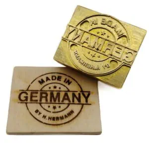 Ein rundes Stempelmotiv, welches in drei Teile gespalten ist. Die Grundformen bestehen aus zwei Kreisen, welche in der Mitte von zwei Linien unterbrochen werden. Das bildet eine Spalte in der der Schriftzug GERMANY seinen Platz findet. Die Buchstaben dieses Wort sind groß und fett dargestellt. Im oberen Bereich der zwei Kreise ist ein gebogenes MADE IN zu lesen, welches sich den zwei Kreisen durch die Biegung anpasst. Im unteren Bereich des inneren Kreises sind jeweils drei Sterne eingebaut, diese finden sich auch im selben Kreisabteil unterhalb von Germany wieder. Im zweiten äußeren Kreis der unteren Hälfte ist ein Schriftzug ebenfalls halbrund gebogen. Geschrieben steht dort BY M. Herrmann, die Art der Darstellung ist kleiner als das Made in, jedoch vom Stil die gleiche Darstellung. Ein gefräst ist das Motiv in einer Messingplatte und wurde auf ein kleines Stück Holz eingebrannt.