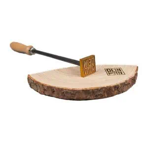Ein Brandstempel mit hellem Holzgriff, anschließender Edelstahlzylinderhalterung und einer Messingplatte, deren Motiv alernativ angepasst werden kann. Das Brenneisen liegt auf einer angeschnittenen Holzscheibe, diese hat ein Brandzeichen der zu sehenden Messingplatte : Dein Motiv - als Text.