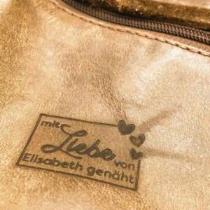 Ein Brandstempel auf dem Material Leder abgebildet. Ein Rahmen, der in der rechts oben von drei Herzen unterbrochen wird. In dem Rechteck befindet sich der Text mit Liebe von Elisabeth genäht. Liebe als Wort wurde in geschwungener Schrift dargestellt.